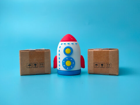 A toy rocket stands centered between two small cardboard delivery boxes on a light blue surface