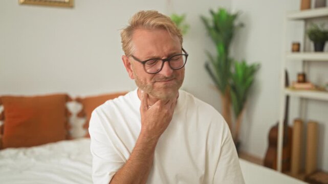 Man clutching throat with hand while seated on bed in a bright bedroom wearing glasses and white tshirt showing pained expression; discomfort.