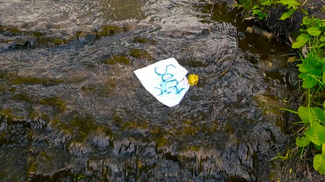 Crumpled white paper with the handwritten word sorry and a sad emoticon floating away on the current of a shallow, rocky river, symbolizing forgiveness, farewell, or letting go of sadness