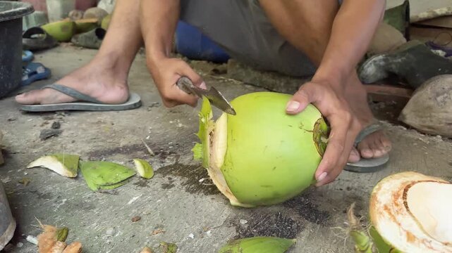 Man Chopping Fresh Green Coconut with a Traditional Machete, A local man wearing flip-flops sits on the ground and uses a large traditional knife to carefully chop open a fresh young green coconut for