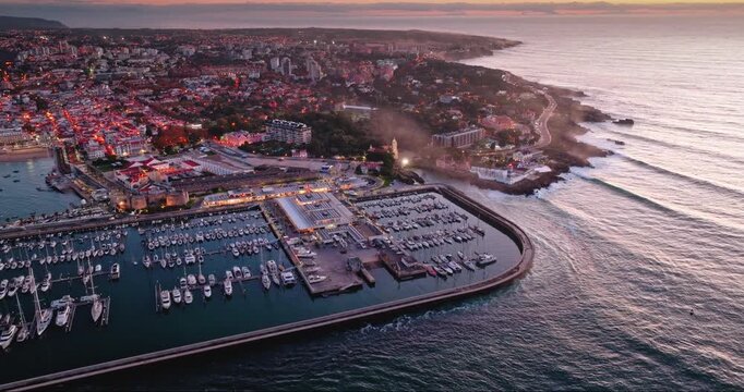 Vibrant Cascais marina in Portugal filled with boats and illuminated coastal city at dusk, Atlantic Ocean coastline and city lights under sunset bright sky. Aerial view drone flight zoom out panorama