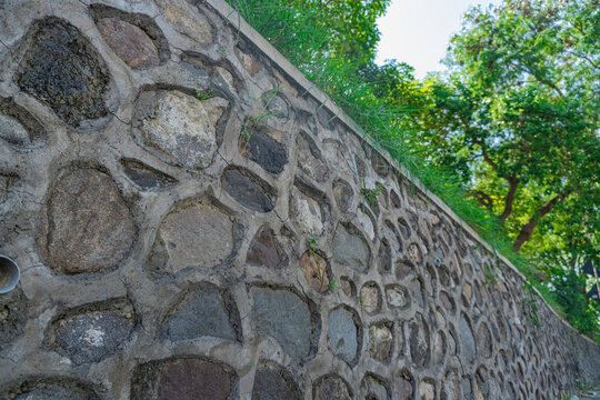 Textured handmade stone retaining wall with concrete mortar and green grass edge under bright daylight trees.