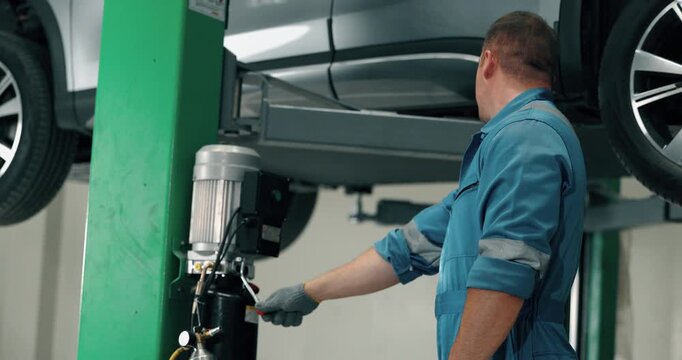 Male mechanic in blue overalls working under car raised on hydraulic lift in auto repair garage