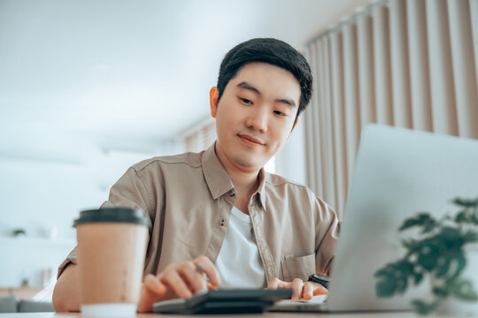 Happy young Asian man working on laptop in living room. Freelance worker male sitting at desk writing notes while watching webinar, studying online