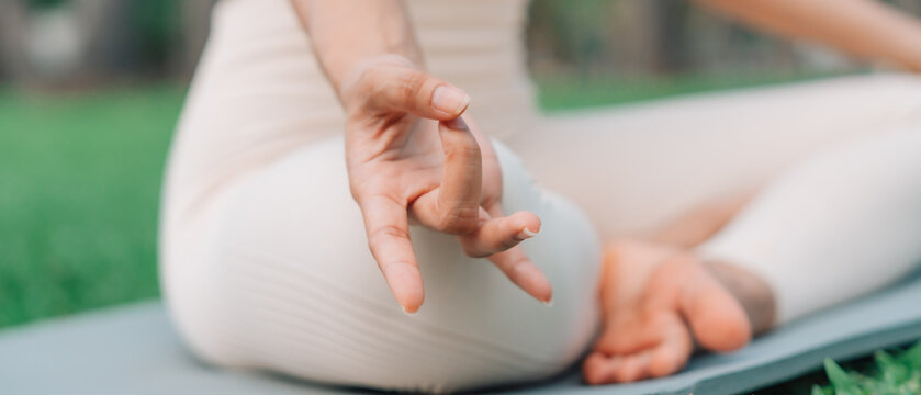 Asian woman meditating in a peaceful park setting, sitting cross-legged on green grass with trees in the background on a sunny day. Concept of mindfulness, self-care, and outdoor relaxation