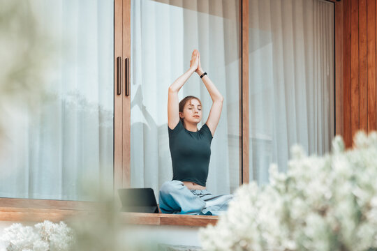 Young woman meditating on a wooden terrace at home, sitting in lotus pose with calm expression. Concept of mindfulness, wellness, and healthy lifestyle in a peaceful modern environment