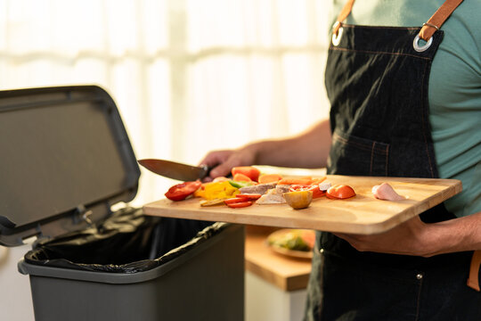 Close up of man in kitchen apron throwing away fresh food scraps into trash bin.