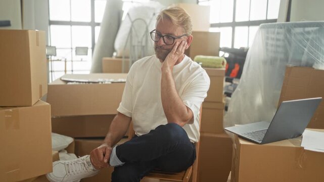 Man seated among packed cardboard boxes touching cheek with hand beside laptop in a building; pensive relocation planning.