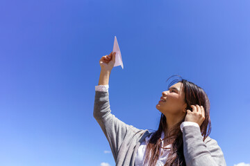 Young Asian woman launching paper airplane towards clear blue sky, expressing future aspirations and dreams © Javier Zayaz