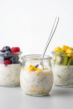 Three vertical glass jars of healthy organic chia seed pudding topped with vibrant fresh mango, kiwi, and mixed berries on a white background, with a silver spoon in the middle jar.