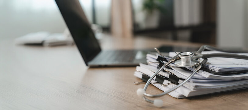 Stethoscope rests on clipped paper documents beside an open laptop on a wooden desk, with wide copy space for medical administration, healthcare records, and presentation messaging.