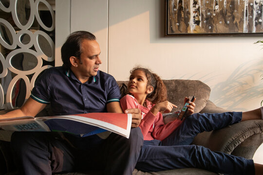 Young girl asking for help from her father while he reads the newspaper at home. 