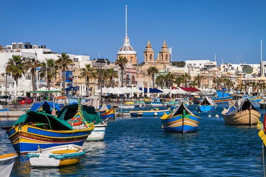 Traditional luzzu boats rest in Marsaxlokk harbor, with palm trees, historic architecture, and church towers beneath a bright blue sky, Malta.