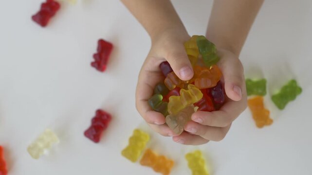Child holds colourful gummy bears with both hands after gathering them from white surface. Close-up overhead composition expresses excitement, playful reward and enjoyment of favourite jelly candy.