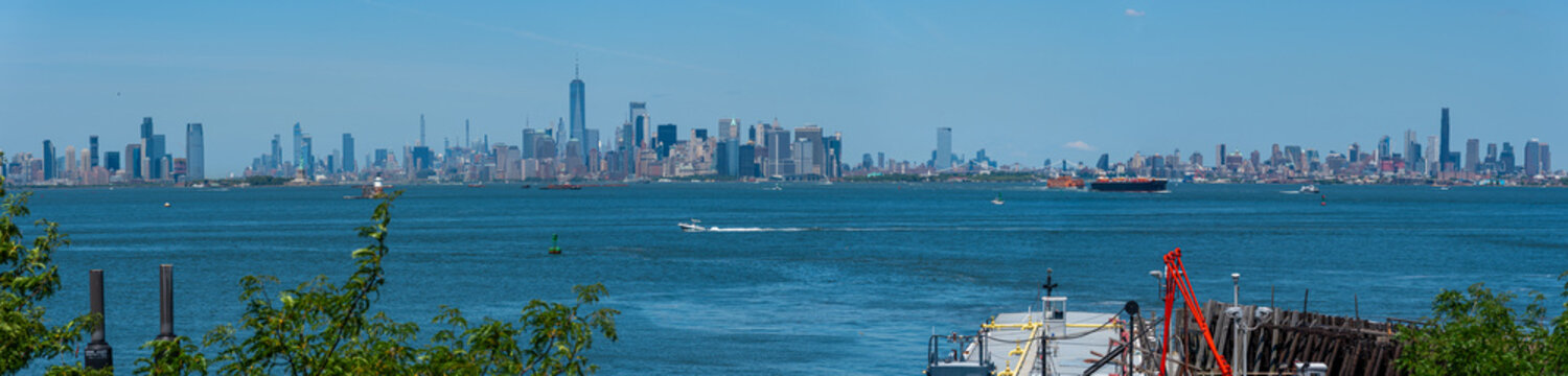A sweeping tri-city panorama: Jersey City, Lower Manhattan, and Brooklyn framed by clear blue skies and the sparkling New York Harbour.