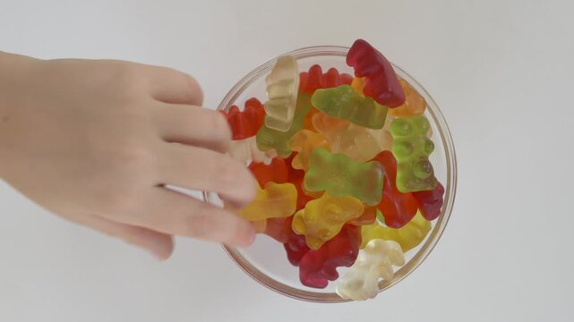 Child quickly grabs colorful gummy bears with both hands from glass bowl on white background. Sugar temptation, impulsive eating, sweet craving and excessive sugar consumption concept.