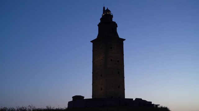Detalle nocturno del faro de la Torre de H&eacute;rcules iluminado, destacando sobre el cielo oscuro y el paisaje costero de A Coru&ntilde;a.