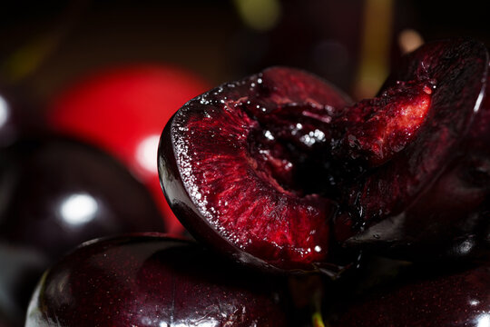 Macro close-up shows sliced dark red cherry with glossy juicy flesh, visible pit cavity, rich texture, and dramatic low light contrast against dark background for fresh food, dessert, and ingredient