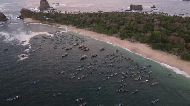 Aerial view of Papuma Beach with numerous fishing boats anchored in the bay near rocky cliffs and lush coastal vegetation in East Java, Indonesia.
