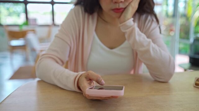 Cropped shot of a bored young Asian woman resting her chin on hand, holding a smartphone while waiting for a message