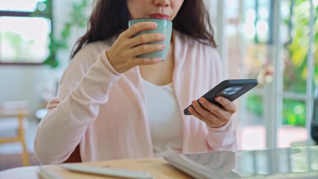 Young woman holding a coffee cup and checking her smartphone at a desk with a laptop and notebook; digital nomad and remote working concept