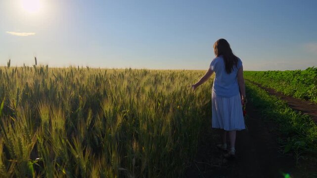 A young teenage girl in a white dress walks along a rural dirt road next to a golden wheat field, gently touching the grain during a warm summer sunset