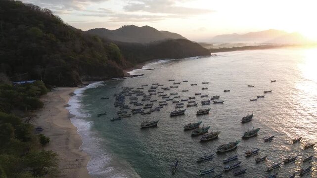 Aerial view of Papuma Beach with numerous fishing boats anchored in the calm bay at sunset, surrounded by lush hills and sandy coastline in East Java, Indonesia.