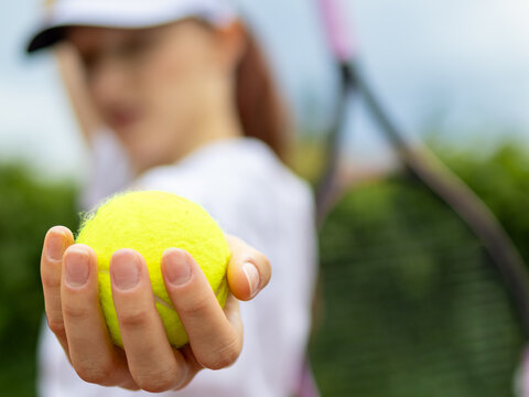 Close-up of a tennis ball held toward the camera by a teenage girl with a racket blurred in the background during outdoor practice. Concept of tennis training, focus, readiness and active lifestyle.