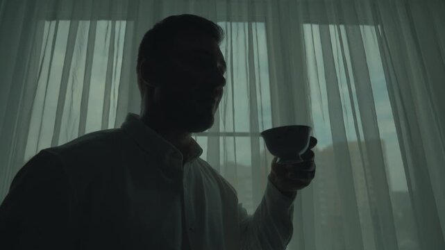 A man in a white shirt is drinking a cup of tea, smiling and enjoying life. The silhouette of a man against the background of the window.