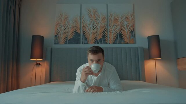 A man in a white shirt drinks a cup of coffee while lying in bed. The concept of a carefree life and enjoyment. When you've earned a lot of money and life is good.