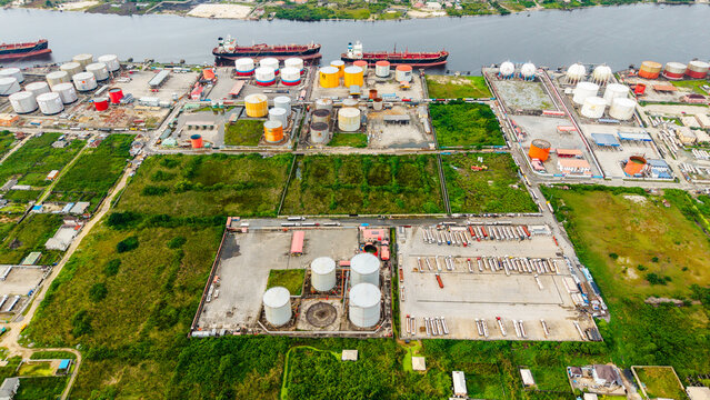 Aerial view of the industrial oil storage facility with large cylindrical tanks, rows of parked tanker trucks, and cargo ships docked along the waterway in Satellite Town, Lagos, Lagos, Nigeria.