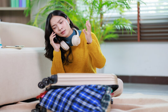 Young asian woman experiencing travel anxiety and frustration, making a distressed phone call while struggling to close an overpacked suitcase before a trip