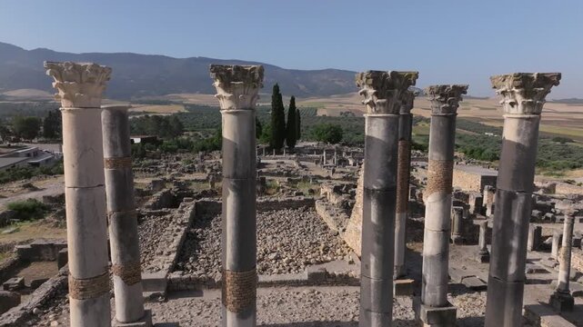 Low-altitude drone view of the monumental Capitol columns at the UNESCO World Heritage-listed Volubilis Archaeological Site in Morocco, overlooking the ancient Roman city and fertile plains.