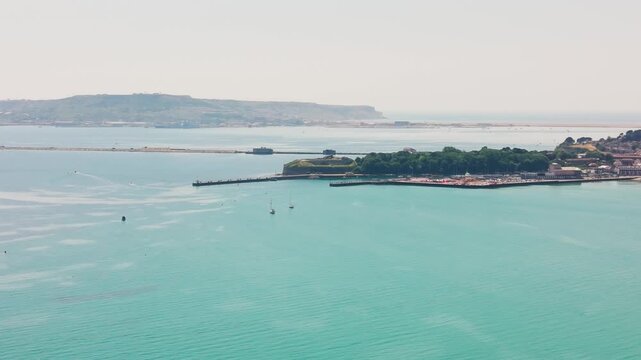 Elevated aerial view across turquoise water at Weymouth Bay, showing Nothe Fort, harbour walls, anchored boats and the Isle of Portland in the distance on a bright summer day.