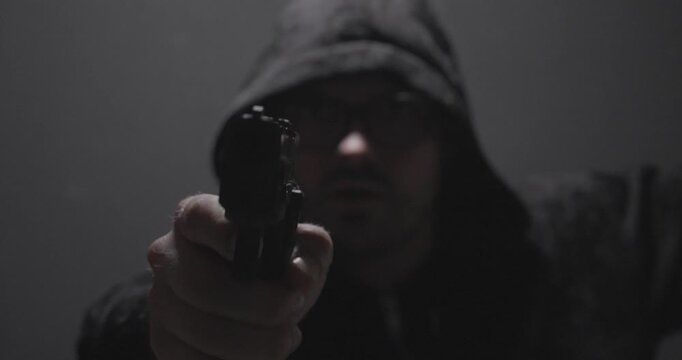 Hooded man aiming a handgun directly at the camera in a dark environment, representing crime, personal threat, armed robbery, danger, and security concepts.