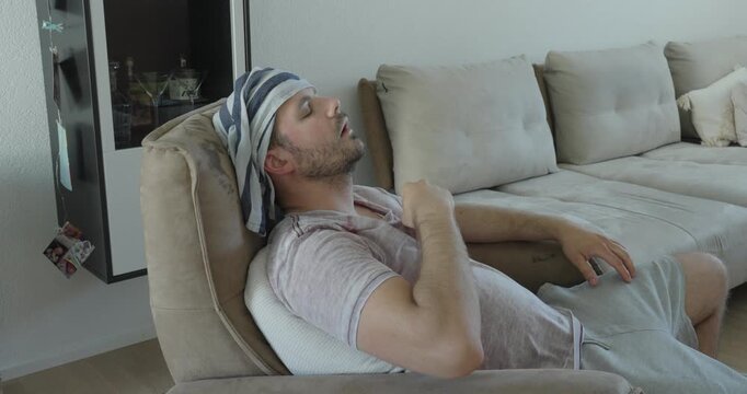 Exhausted man resting on a sofa with a wet towel on his head during an extreme heatwave, trying to cool down from high indoor temperatures.