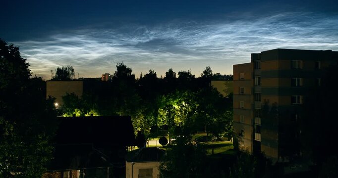 Rare noctilucent clouds glowing above residential buildings and trees at night. Luminous night clouds create a striking atmospheric display over the illuminated urban landscape.
