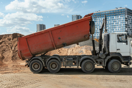 Heavy tipper truck with raised orange dumper body operating at modern city construction site. This commercial image is ideal for real estate development, logistics and industrial machinery