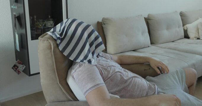 Exhausted man placing a wet towel over his face while resting on a sofa during an extreme heatwave, trying to cool down and relieve heat stress.