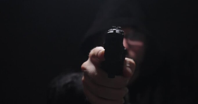 Hooded man aiming a handgun directly toward the camera in a dark setting, representing crime, personal threat, danger, armed robbery, and security concepts.