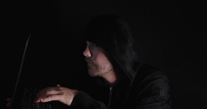 Hooded man pointing at a laptop screen in a dark room, representing hacking, cybersecurity, cybercrime, and digital security concepts.