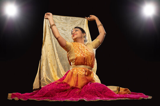 Kathak dancer sitting gracefully and performing in front of a dark background. 
