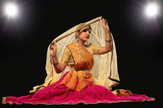 Kathak dancer sitting gracefully and performing in front of a dark background. 