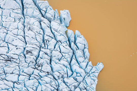 Aerial view of Jokulsarlon glacier edge with deep blue crevasses and textured ice formations meeting the calm glacial lagoon water Sveitarfelagid Hornafjordur, Iceland.