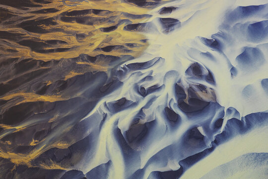 Aerial view of a glacial river in Iceland, showing intricate patterns of blue water flowing through dark volcanic sand, creating a striking contrast. Selfoss, Iceland.