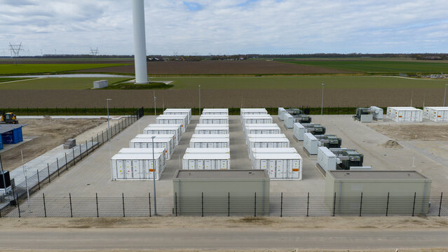 Aerial view of a battery energy storage system with rows of white containers and electrical equipment in a fenced facility near a wind turbine in Dronten, Flevoland, Netherlands.