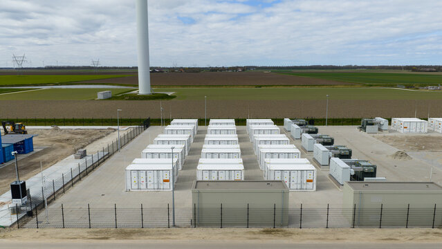 Aerial view of a battery energy storage system with rows of white containers on a paved industrial site near a wind turbine in Dronten, Flevoland, Netherlands.