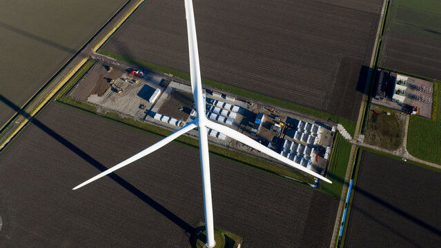 Aerial view of a large white wind turbine standing next to a battery energy storage system facility surrounded by vast agricultural fields in Dronten, Flevoland, Netherlands.