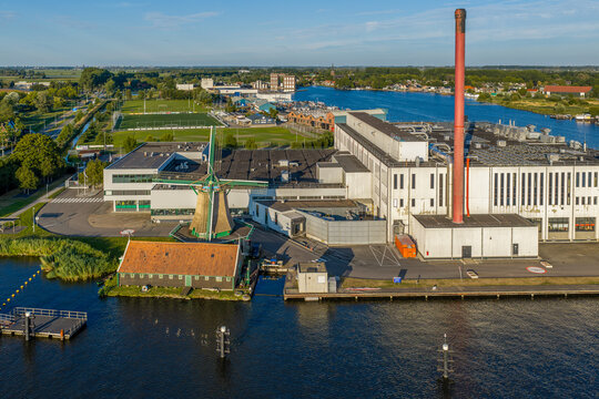 Aerial view of the Duyvis Production B.V. industrial facility with a traditional windmill, modern factory buildings, and a tall chimney near the river in Zaandam, Noord-Holland, Netherlands.