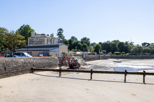 Excavator carrying out beach maintenance and coastal engineering work at low tide on the Isle of Wight with construction machinery reshaping sand beside a stone seawall beneath a clear blue summer sky